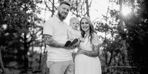 Family portrait in a forest, with a tattooed father, mother holding a baby, and warm lighting.